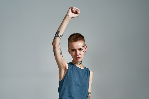 Disgruntled tattooed young caucasian woman protester with piercing looking at camera while posing with raised arm isolated over gray background. Front view. Horizontal shot