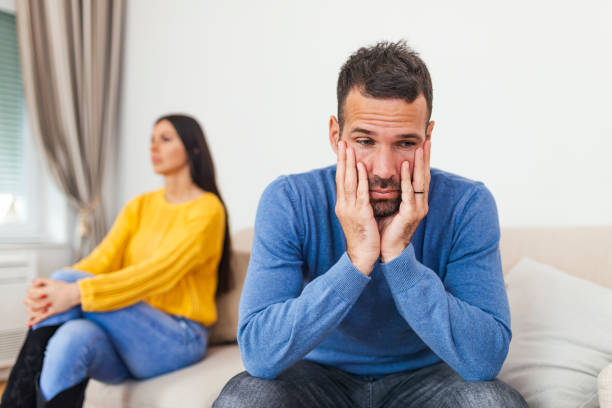istock Young man sitting on a couch is having an argument with his girlfriend who has her head turned away. Couple sitting on the couch not speaking after a fight at home in the living room 1303618533