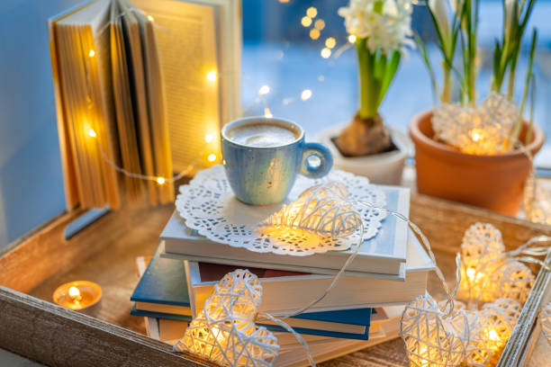 Coffee cup, open book, spring flowers, sparkle lighting garland on window sill on blue blurred background. Concept warm and cozy home, reading books, hygge, evening rest and comfort leisure