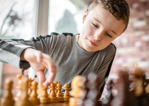 Cute young happy boy chess genius concentrating playing game of strategy moving piece sitting at wooden table with chessboard on bright sunny summer day