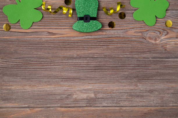 istock Patricks day background with copy space. Green clover leaves and hat with golden glitters coins on the brown wooden table, flat lay, top border 1303622232