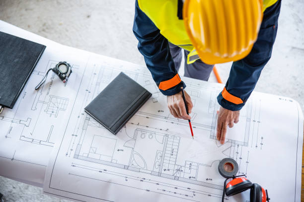 istock Overhead view of construction worker in hard hat inspecting building blueprint plans on the table 1303623675