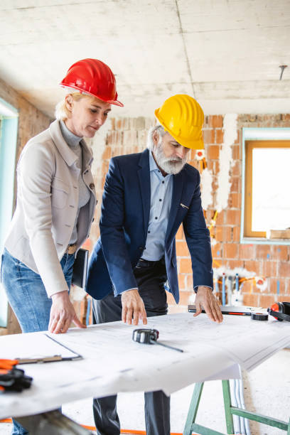 istock Male construction site supervisor and female client in hard hats analyzing building blueprints inside newly built structure 1303623676