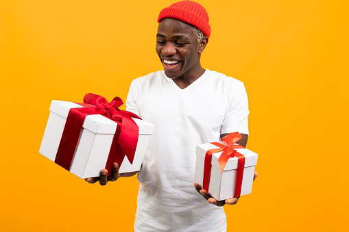 funny american man with a smile in a white t-shirt holds two gift boxes with red ribbon on a yellow studio background.