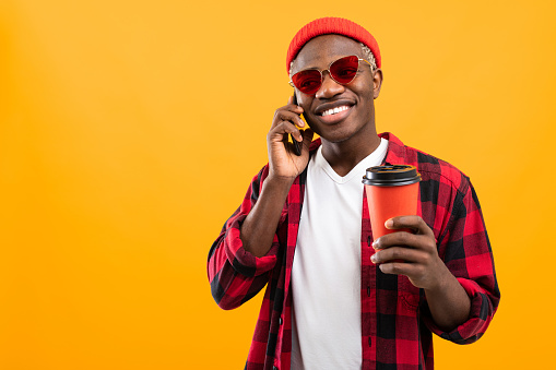 handsome black american man talking on the phone while holding a take-away coffee glass on yellow studio background with copy space.