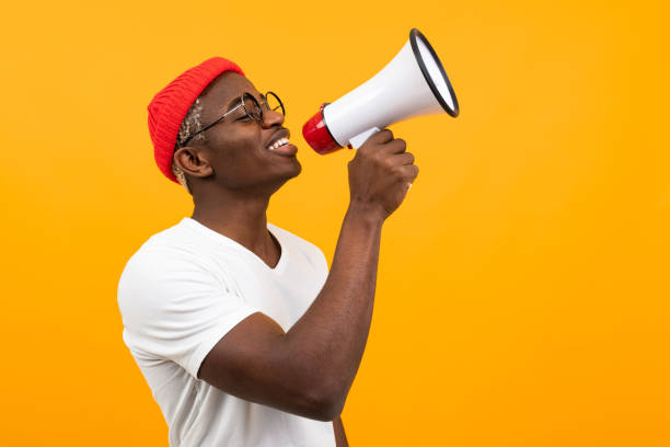 black handsome smiling american man in white t-shirt speaks news through a megaphone on isolated orange background with copy space black handsome smiling american man in white t-shirt speaks news through a megaphone on isolated orange background with copy space. black guy with blonde hair stock pictures, royalty-free photos & images