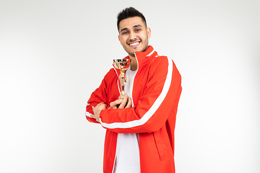 man in a red tracksuit shows off the winner's gold cup on a white background.