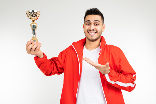 man in a red tracksuit shows off the winner's gold cup on a white background.