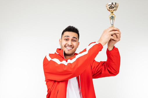 man in a red tracksuit holds a winner's gold cup raised up on a white background.