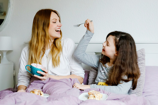 Lovely young mother and cute school kid girl cuddling together in bed in morning. Happy family of young single woman and beautiful daughter having breakfast, eating on mother's day