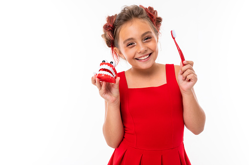 Portrait of little caucasian girl with fair hair and pretty face in red dress smiles and rejoices with tooth brush