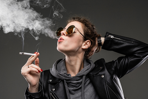 Portrait of beautiful caucasian female in grey hoodie, black jacket, black sunglasses smokes