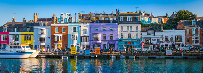 Colourful cottages, fishing boats and yachts around the harbour as holiday makers and locals enjoy the warm summer evening outside waterfront pubs and restaurants at the popular seaside resort town of Weymouth, Dorset.