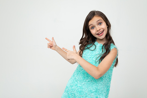 smiling teenage girl in a blue dress points her hands towards empty space on a white studio background.