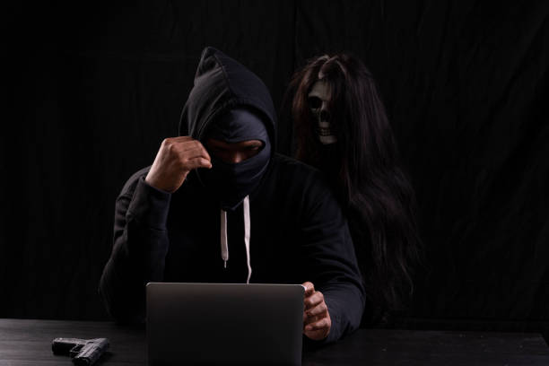 istock Mysterious man in the hood with skull inside using laptop on table with gun on isolated black background. Hacker male hoodie anonymous use networking with ghost. Halloween and technology concept. 1303640142
