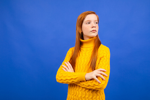 serious charming european red-haired teenager girl in a yellow sweater on a blue studio background.