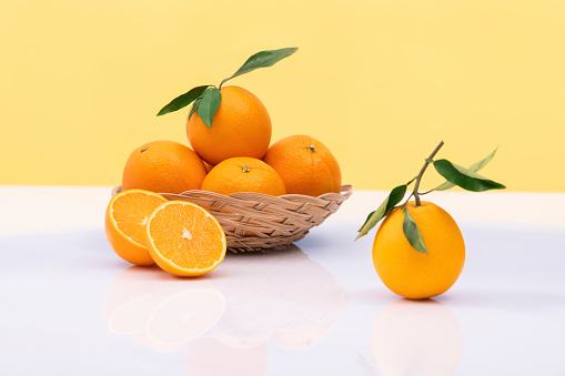 Fresh oranges in a wicker basket with leaves on a glass table on an yellow background. selective focus. vitamin and healthy food concept.