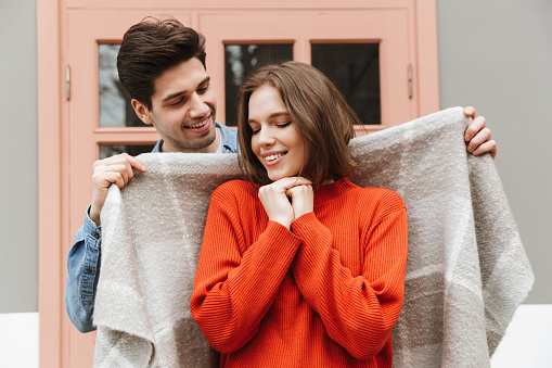 Photo of smiling couple standing outside and man wrapping his woman in warm blanket