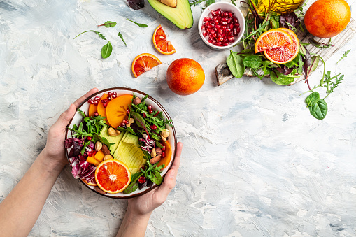 Girls' hands holding vegan, detox Buddha bowl with avocado, persimmon, blood orange, nuts, spinach, arugula and pomegranate on a light background, top view.