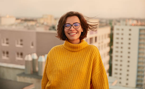 Positive young lady on roof Happy young female in trendy sweater and glasses smiling and looking at camera while standing on roof and resting in weekend evening in city town of hope stock pictures, royalty-free photos & images