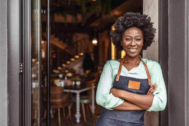 I sacrificed a lot for this startup to come true Cheerful female café owner standing at the door with her arms crossed. African woman in apron standing with her arms crossed small business owner stock pictures, royalty-free photos & images