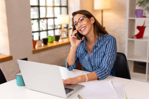 istock Cheerful businesswoman using laptop while talking on mobile phone in the office 1303652873