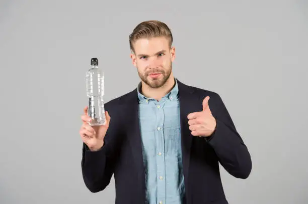 Photo of Bearded man show thumbs up to bottle of water. Thirsty businessman with beard in blue shirt and coat. Thirst and dehydration concept. Drinking water for health