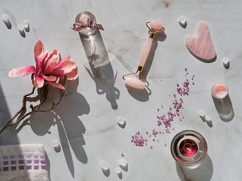 Crystal rose quartz facial roller and Gua sha stone for beauty facial massage therapy, Flat lay on marble table with magnolia flowers. Long shadows and candle. Essential oil bottle.