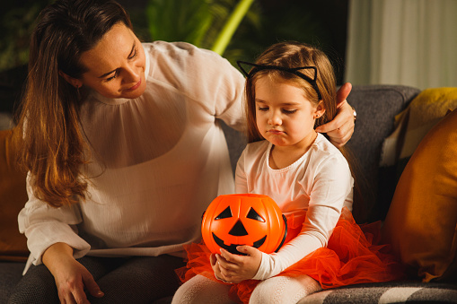 Front view of caring mother consoling her sad little daughter who is holding an empty Jack o' lantern bucket with no Halloween candy.