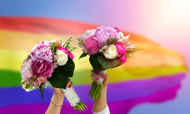 Lesbians in wedding holding a bouquet Lesbians in wedding holding a bouquet of flowers celebrating the wedding against the background of the LGBT flag civil partnership stock pictures, royalty-free photos & images