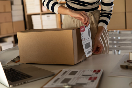 Midsection of unrecognizable female entrepreneur applying a label on a cardboard box with customer's order that she is preparing for shipping.