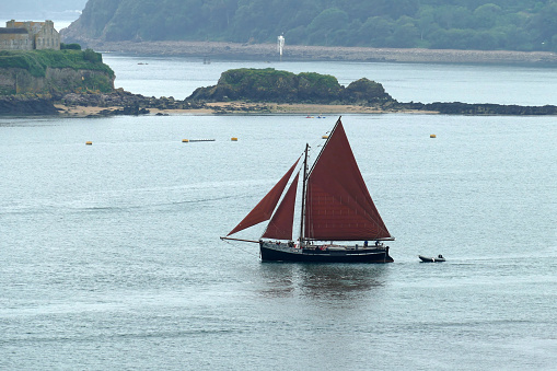 Plymouth England.  Small schooner with red sails passing Drake's island in Plymouth Sound heading out to sea.