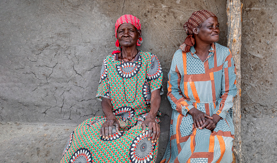 two elderly woman sitting next to the house wall in the yard in an African village