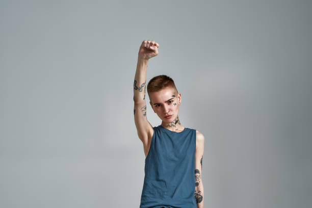 istock Tattooed young caucasian woman protester with piercing looking at camera while posing with raised fist isolated over gray background 1303668171