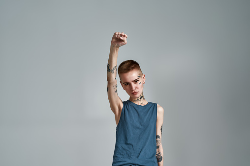 Tattooed young caucasian woman protester with piercing looking at camera while posing with raised fist isolated over gray background. Front view. Horizontal shot