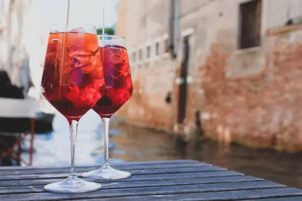 Photo of Two glasses of Spritz Veneziano cocktail served near the Venetian canal.  Popular italian summer aperitif drink. Copy space. Venice background.