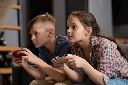 Two cute serious siblings sitting on couch in front of tv set and playing video game in living-room during vacations or on weekend