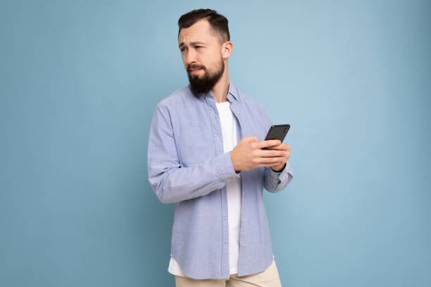 istock Thoughtful handsome young man isolated over background wall wearing everyday clothes holding and using mobile phone writing sms looking to the side and thinking 1303677646