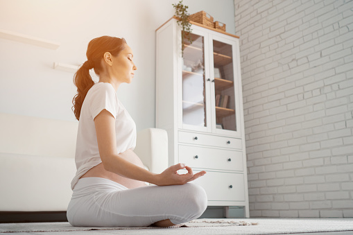 Beautiful pregnant woman with large bare tummy meditates sitting in lotus position at home side view