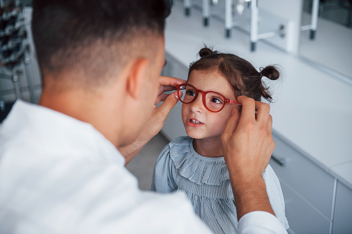 Joven pediatra con bata blanca ayuda a conseguir nuevos anteojos para niña photo
