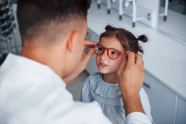 Photo of Young pediatrician in white coat helps to get new glasses for little girl