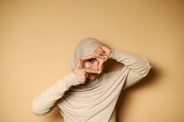 A muslim woman in hijab looks at the camera through her fingers framing her face standing on beige background with copy space Beautiful muslim women in hijab making a frame with fingers and looking through it. Portrait on beige background with copy space. Facial emotions. person holding frame over face stock pictures, royalty-free photos & images