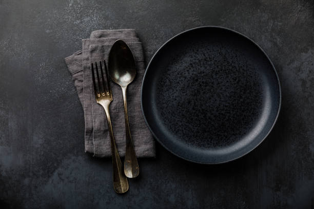 istock Table setting with empty black plate, fork and spoon cutlery and linen napkin on dark table 1303681661