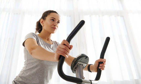 Attractive fit woman exercising on a spin bike at home on a beautiful sunny day. Cardio workout. Fitness at home. Close-up