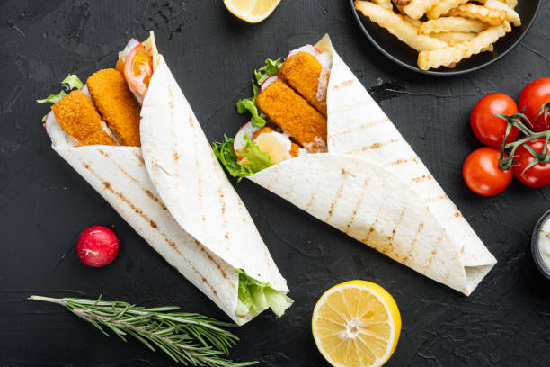 istock Tortilla roll with fish fingers, cheese and vegetables, on black background, top view flat lay 1303685724