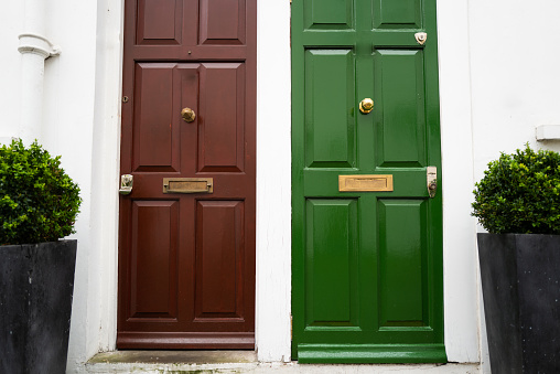 Color image depicting the exterior architecture of terraced residential houses in the borough of Kensington and Chelsea, an affluent area of London, UK. Two doors, one green and one dark red, the entrances to the houses, are side by side.