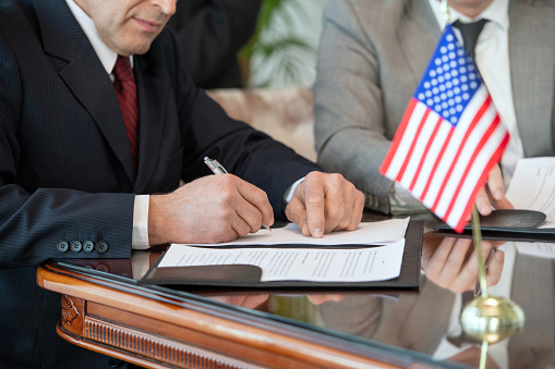 Mature American delegate in formalwear signing contract of business partnership with foreign partner while sitting by table in boardroom