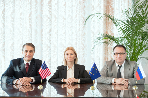 Row of business partners or delegates from EU, USA and Russia sitting by table in front of camera during working meeting in boardroom