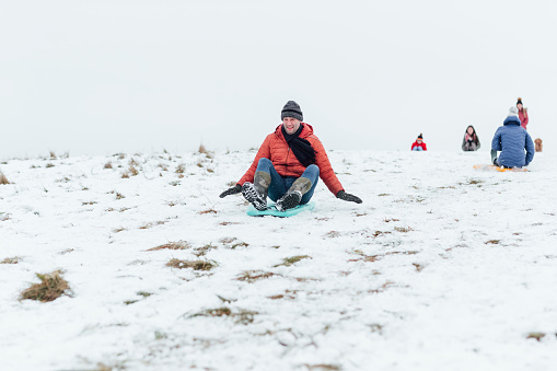 A mature caucasian man wearing warm winter clothing and accessories in a snowy, rural, outdoor setting. He is having fun sledding down a snowy hill on a winters day.