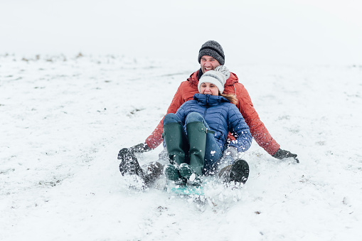 A mature caucasian couple wearing warm winter clothing and accessories in a snowy, rural, outdoor setting. They are sledding down a snowy hill on a winters day.